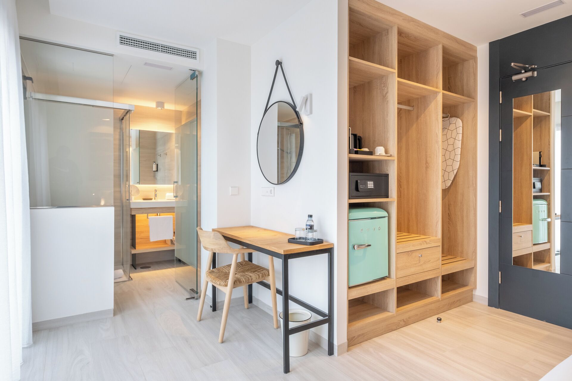 Una habitación de hotel moderna con kitchenette, una zona de comedor con mesa y sillas, un armario con varios compartimentos y un espejo en la pared.