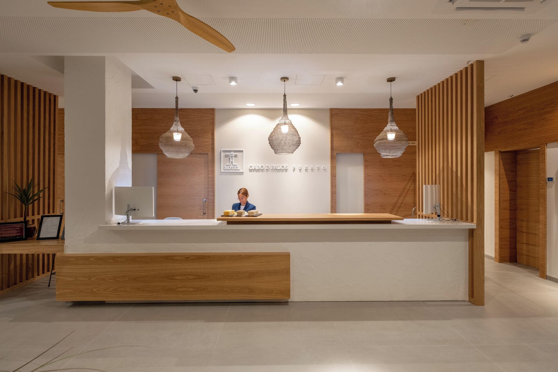 Un comptoir d’accueil d’hôtel avec un réceptionniste, des panneaux de bois et des luminaires suspendus.