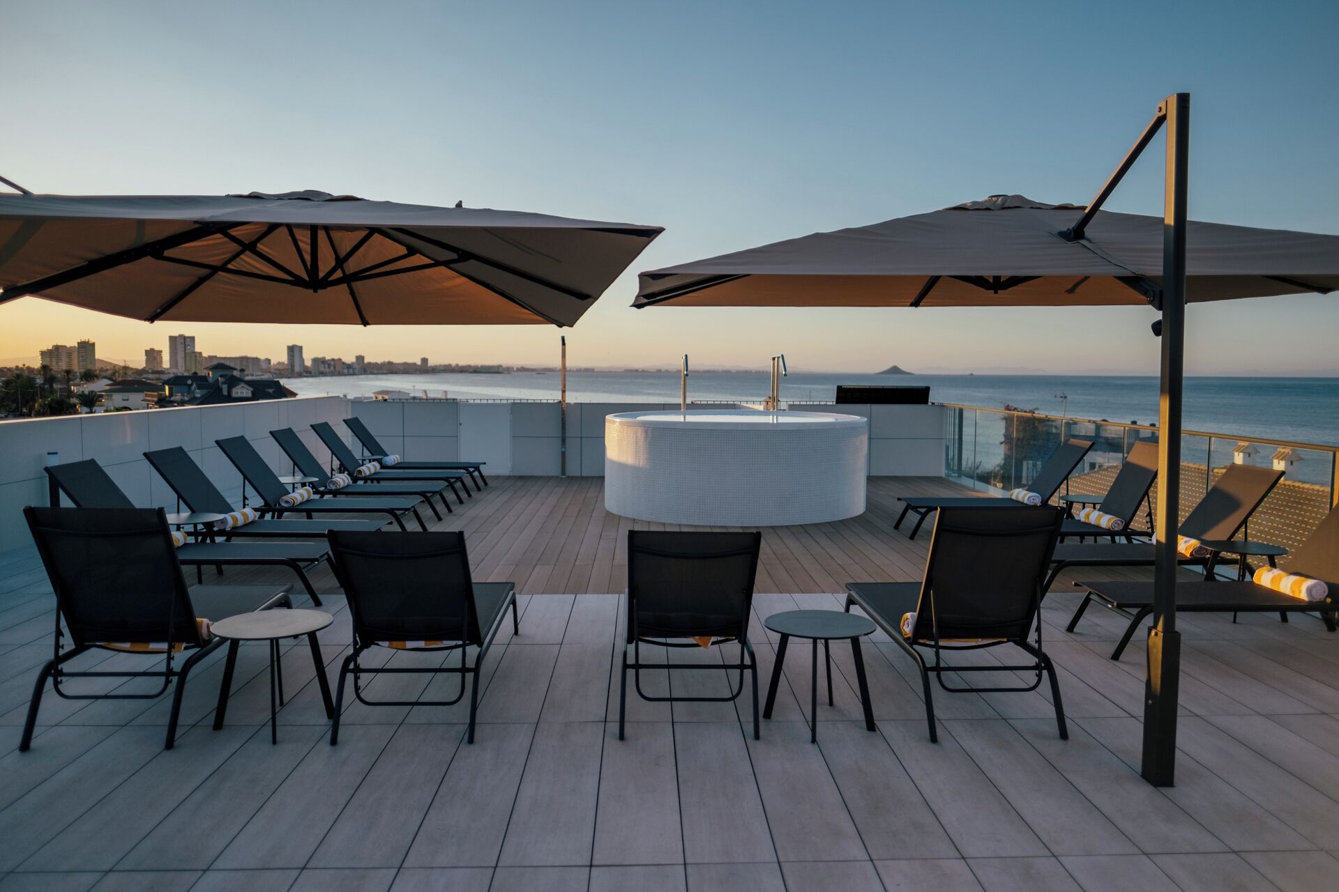 A rooftop terrace with lounge chairs and umbrellas, overlooking a cityscape and the ocean.