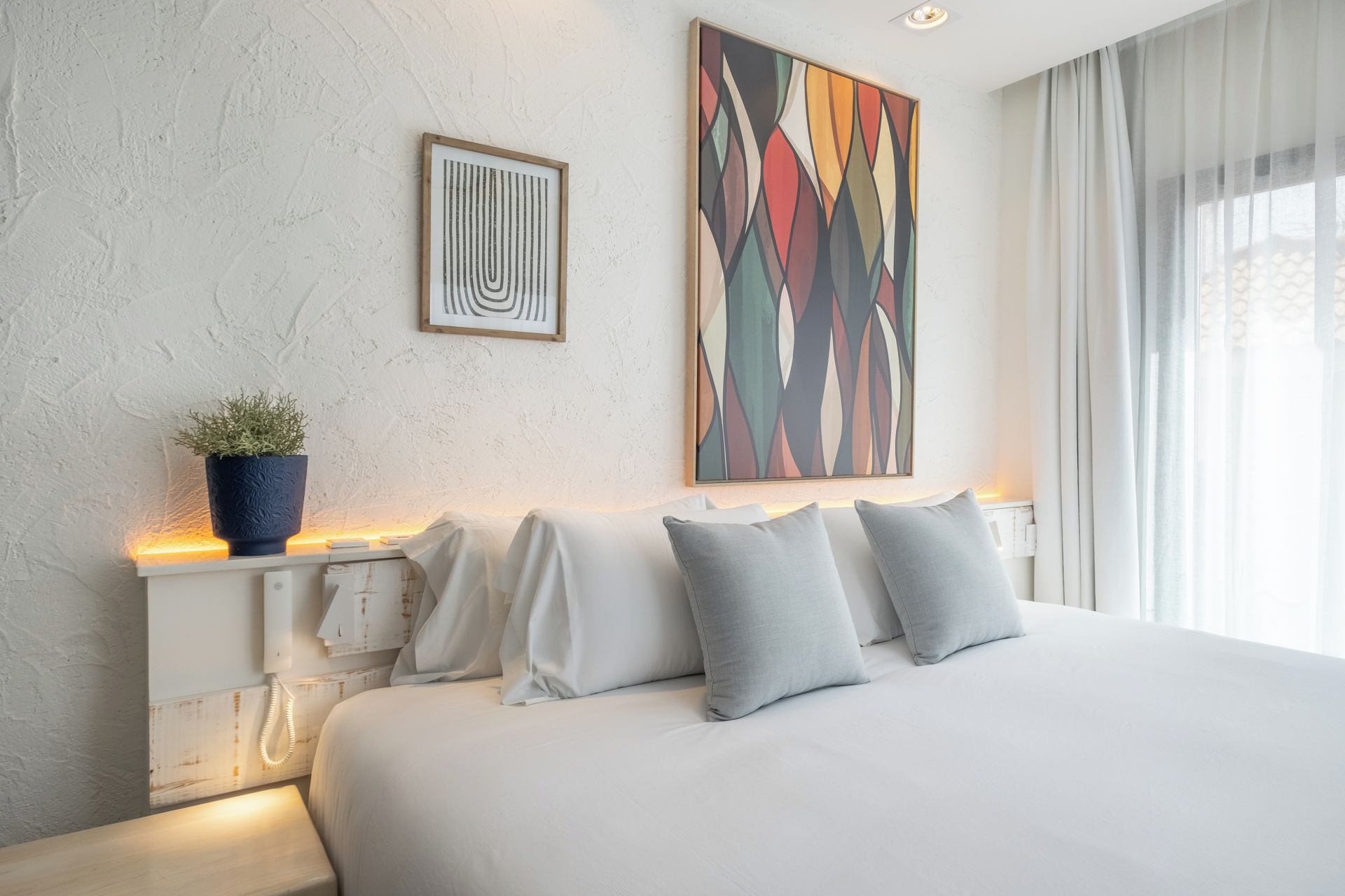 Habitació d'hotel moderna amb un llit gran, una planta en test i dues obres d'art a la paret.