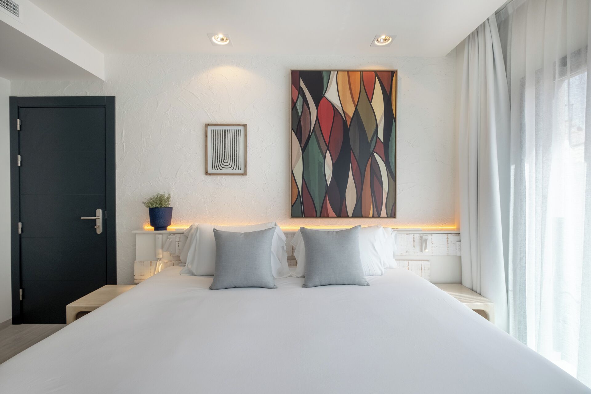 A modern hotel room with a large bed, a dark door, a potted plant, a colorful abstract painting, and a view of the outdoors.