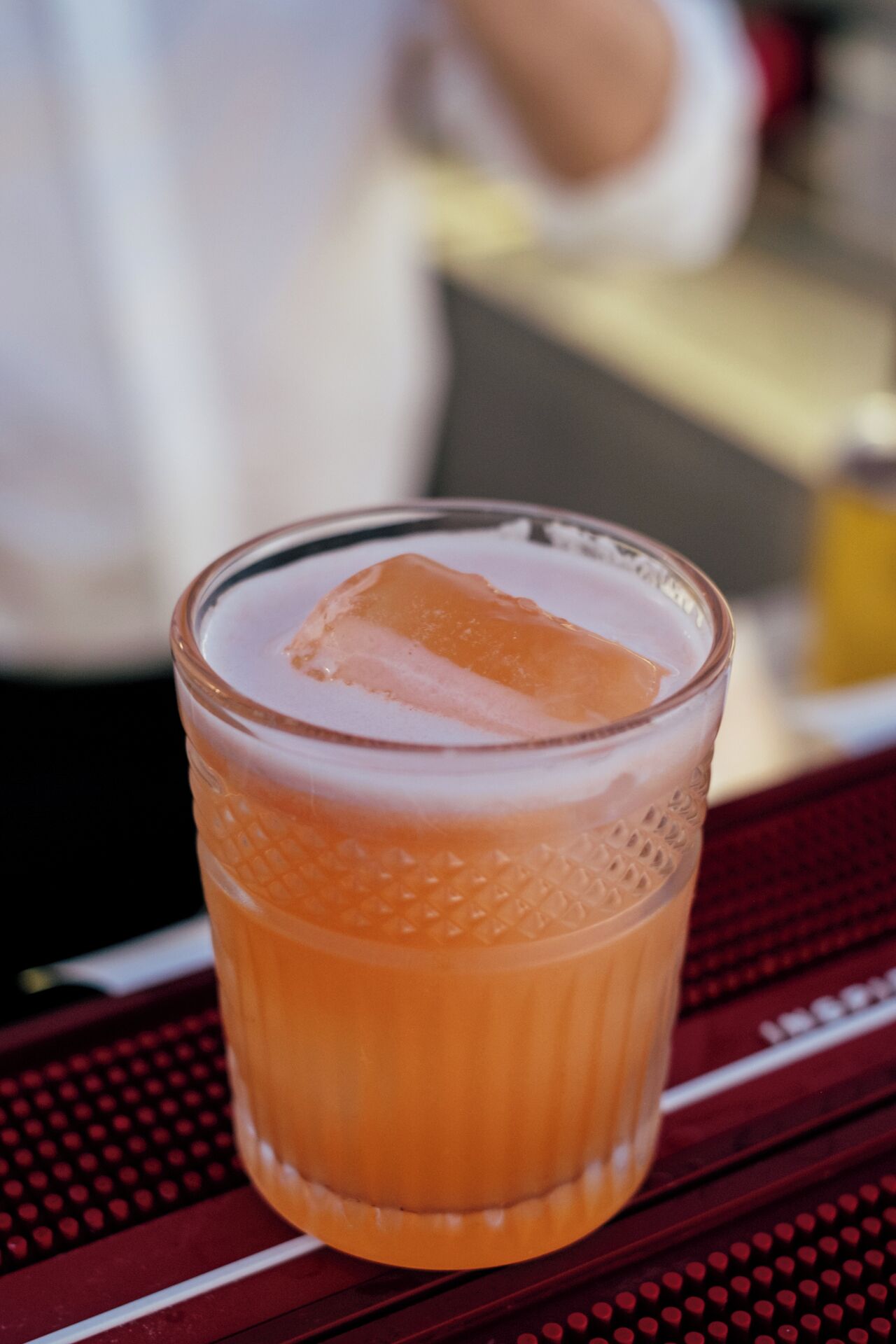 A glass with a beverage and a cube of ice on a table.