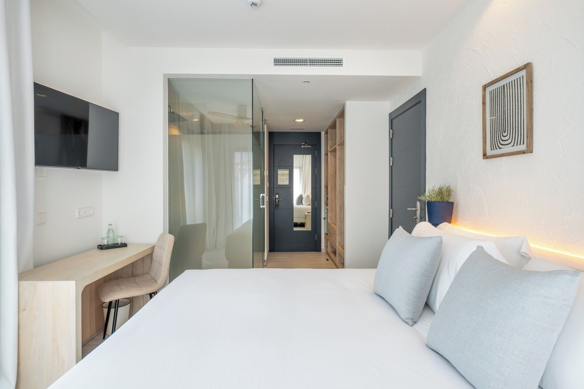 Habitación de hotel moderna con una cama grande, un escritorio y una silla. Hay un televisor montado en la pared, una planta en maceta y una obra de arte enmarcada.