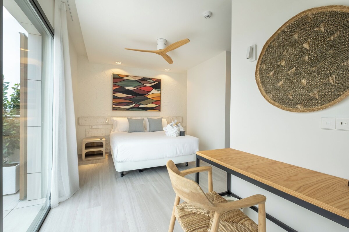 A modern hotel room with a bed, a desk, a chair, a ceiling fan, and a large abstract painting.