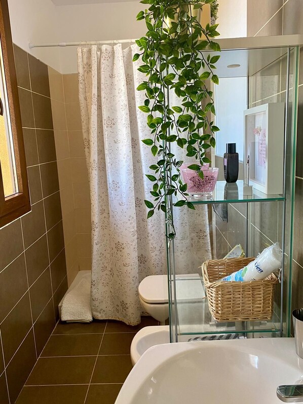 A bathroom with a shower curtain, a glass shelf with toiletries, a wicker basket, and a white bathtub.