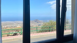 A room with large windows offering a view of a coastal landscape, including a mountain and a road with a green railing. - Casa Amplia y Espaciosa Para Descubrir un Tenerife Auténtico y Local (San Miguel)
