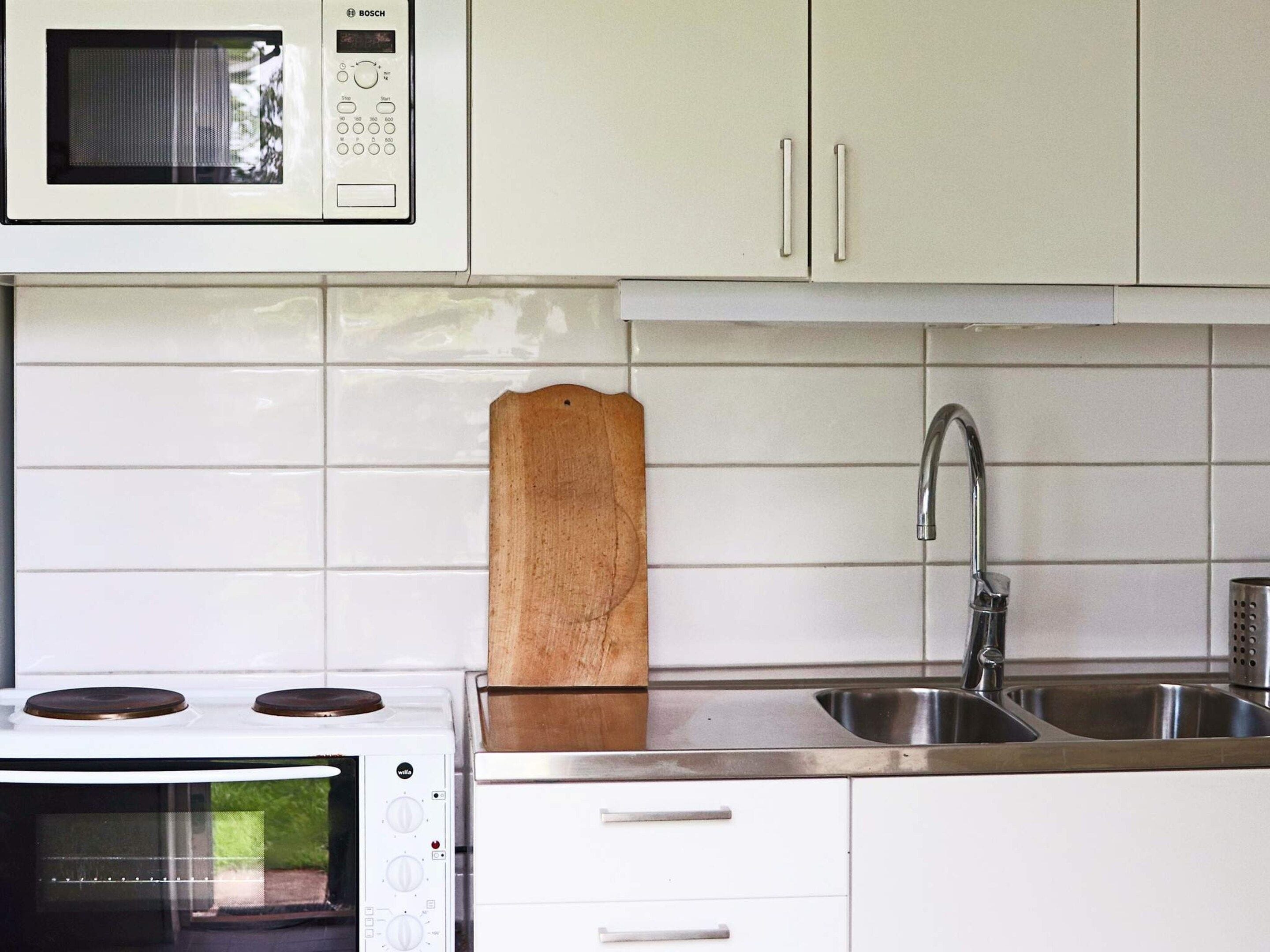 A kitchen with white cabinets, a microwave, a stove, a wooden cutting board, and a stainless steel sink.