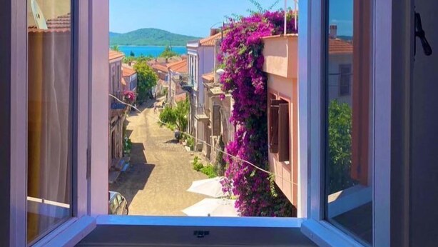 An open window with a view of a narrow street lined with buildings, some with balconies adorned with purple flowers. - Cunda Bebekli Ev (Ayvalik)