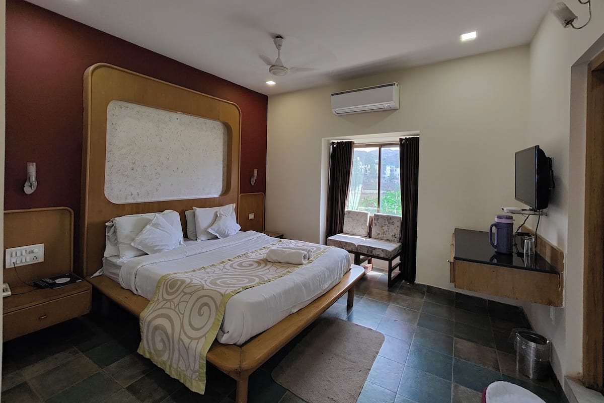 A hotel room with a large bed, a television, a window with curtains, and an air conditioning unit.