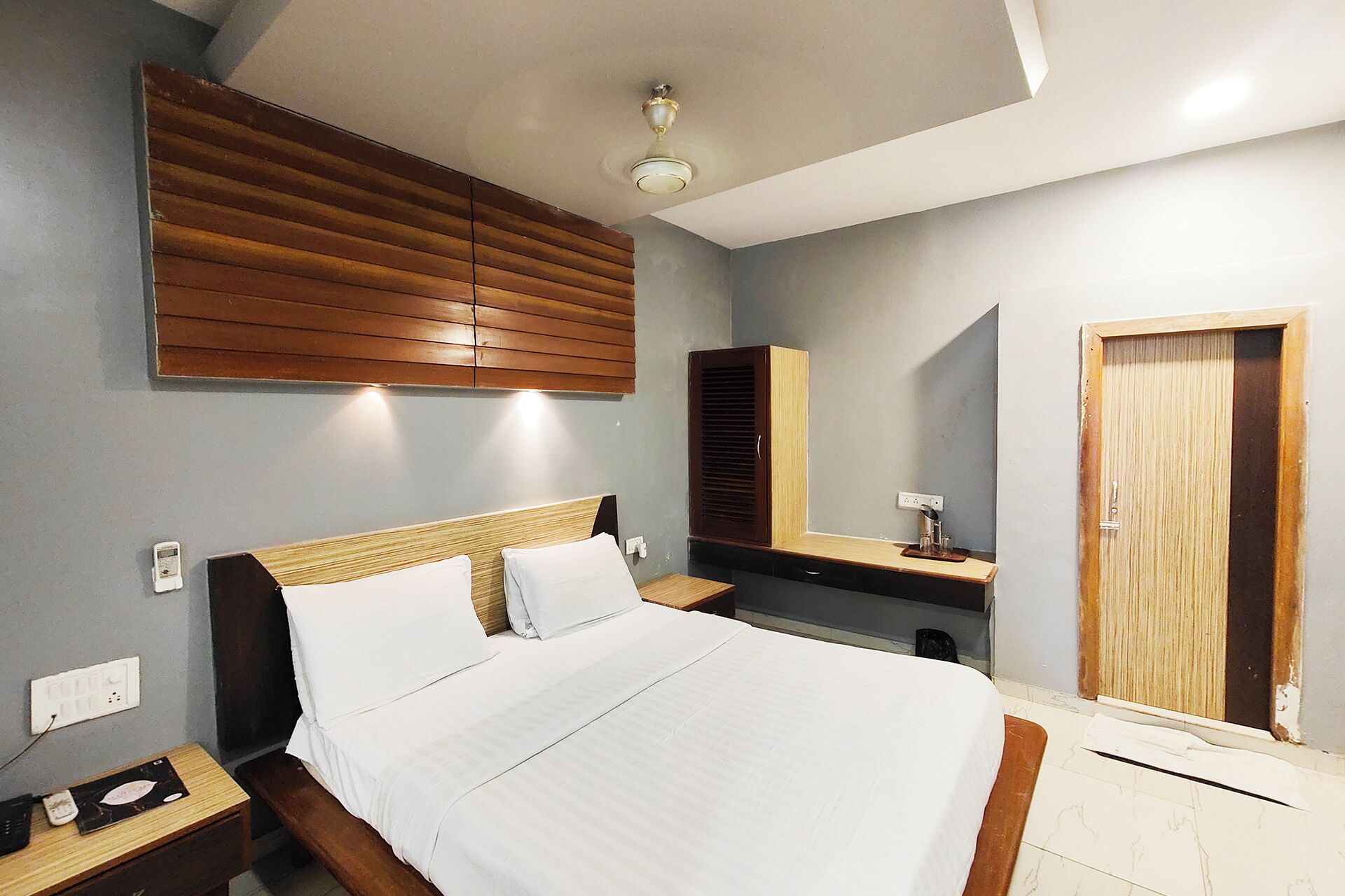 A hotel room with a bed, wooden headboard, bedside tables, a nightstand, a mirror, a wardrobe, and a ceiling fan.