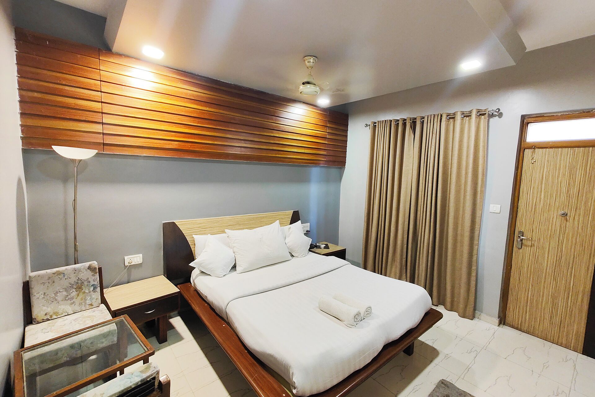 A bedroom with a wooden headboard, a bed with white linens, a bedside table, a nightstand, a floor lamp, and a door with curtains.