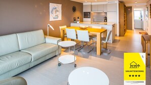 Eine moderne Wohnung mit Wohnzimmer, Essbereich und Küche. Es gibt ein Sofa, einen Esstisch, Stühle und eine Küchenarbeitsplatte.