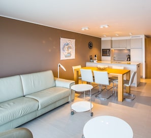 Eine moderne Wohnung mit Wohnzimmer, Essbereich und Küche. Es gibt ein Sofa, einen Esstisch, Stühle und eine Küchenarbeitsplatte.