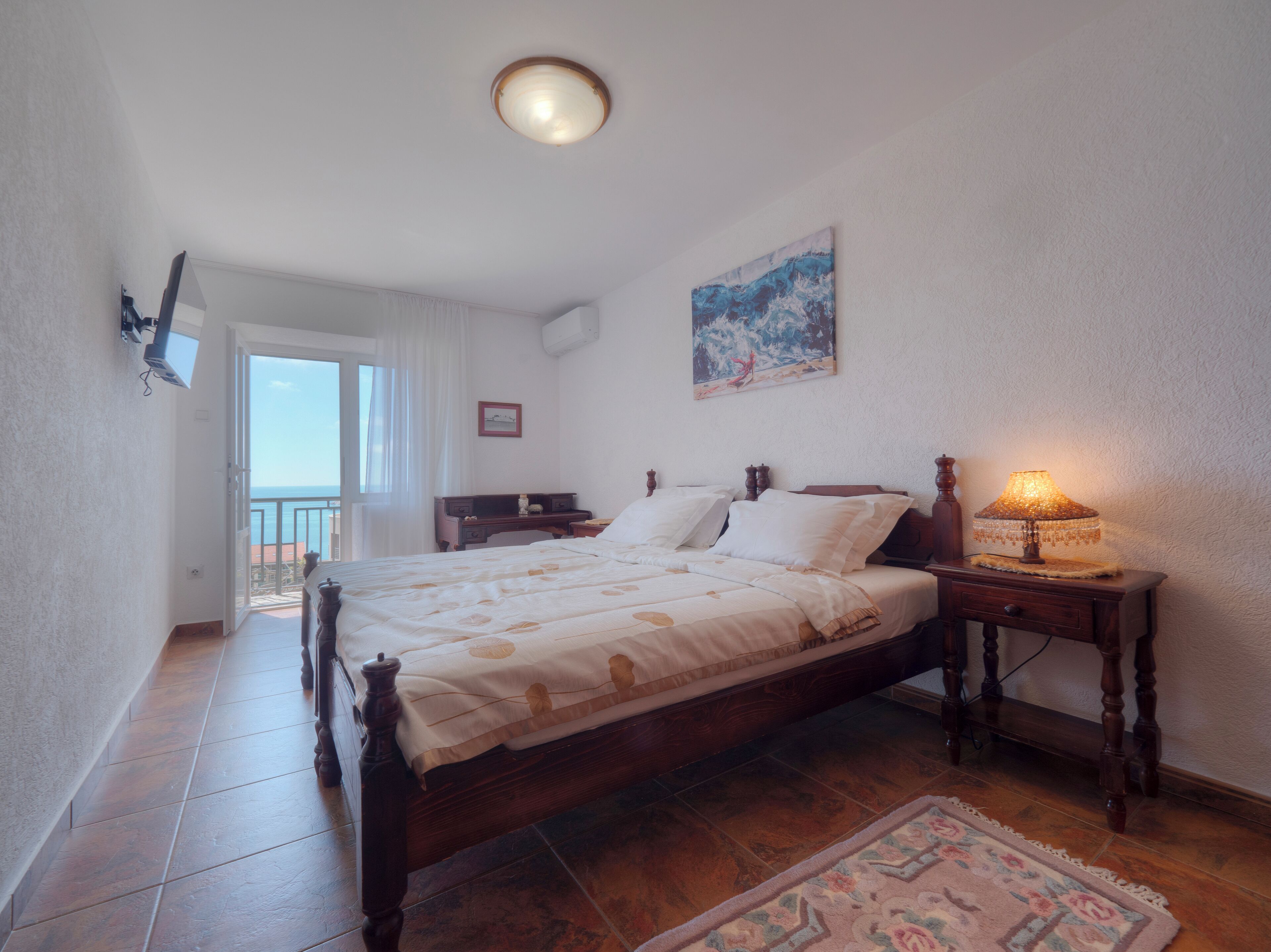 Une chambre avec un lit en bois, une table de chevet avec une lampe, un tableau au mur et une vue sur la mer par la fenêtre.