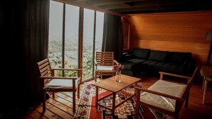 A cozy room with a large window, a black sofa, wooden chairs, a small table, and a patterned rug. - Ayliya Bungalow (Akçaabat)