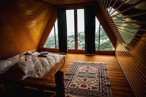 A cozy bedroom with a wooden sloped ceiling, a bed with pillows, a rug, and large windows offering a view of the outdoors. - Ayliya Bungalow (Akçaabat)