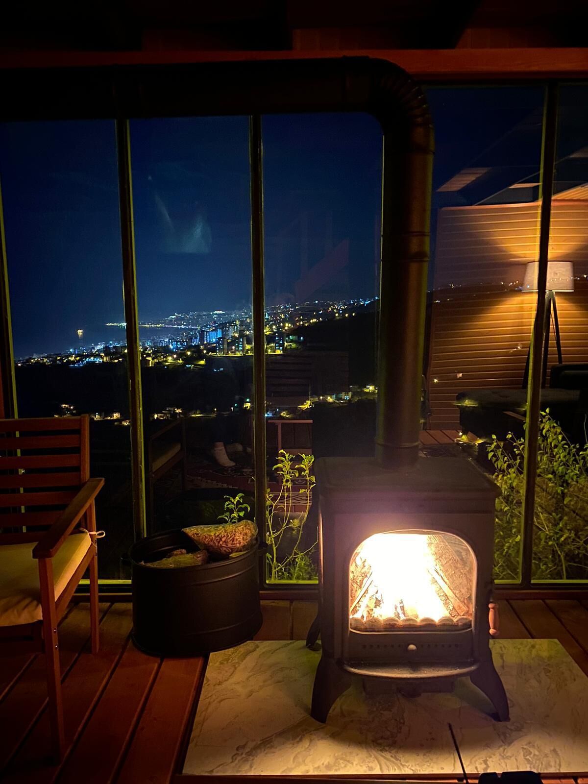 A cozy room with a wood-burning stove, a comfortable chair, and a large window offering a view of a cityscape at night.