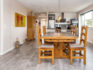 A dining area with a wooden table and chairs, a kitchen in the background, and a large window.