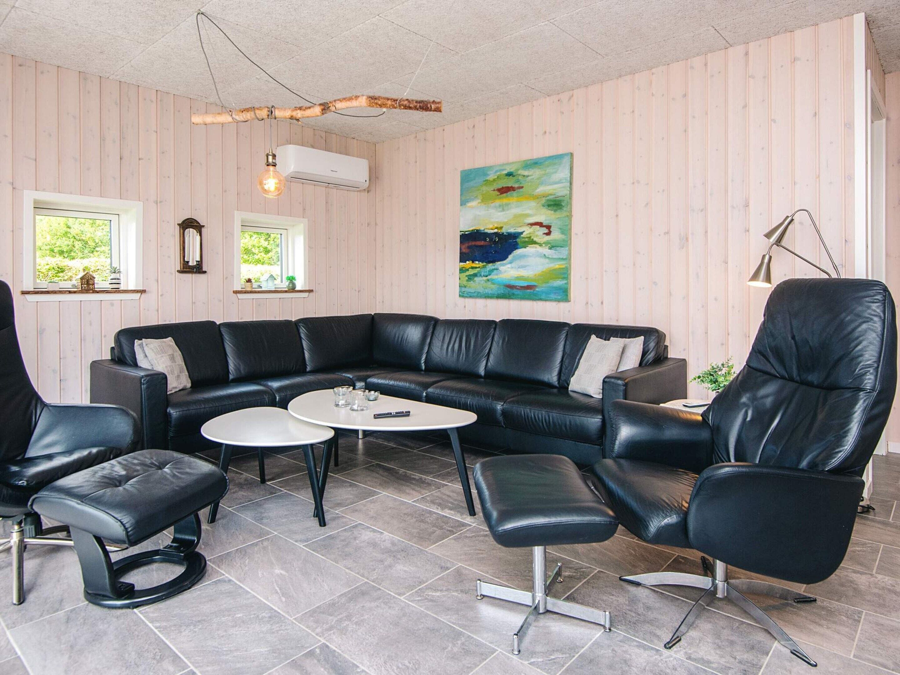 Un salon moderne avec un canapé d’angle en cuir noir, une table basse ronde et des fauteuils assortis en cuir. Un tableau orne le mur, une fenêtre offre une vue sur la verdure et un appareil de climatisation est installé.