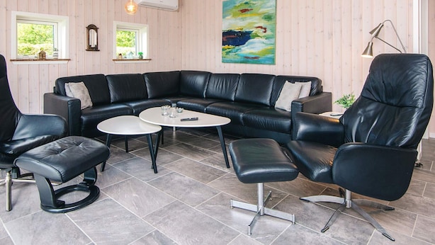A modern living room with a black leather sectional sofa, a round coffee table, and matching leather chairs. There is a painting on the wall, a window with a view of greenery, and an air conditioning unit. - 5 Star Holiday Home in Hemmet (Hemmet)