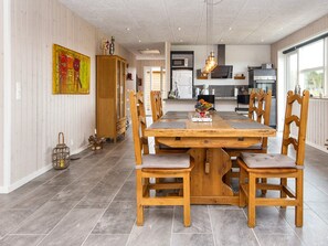 A dining area with a wooden table and chairs, a kitchen in the background, and a large window.
