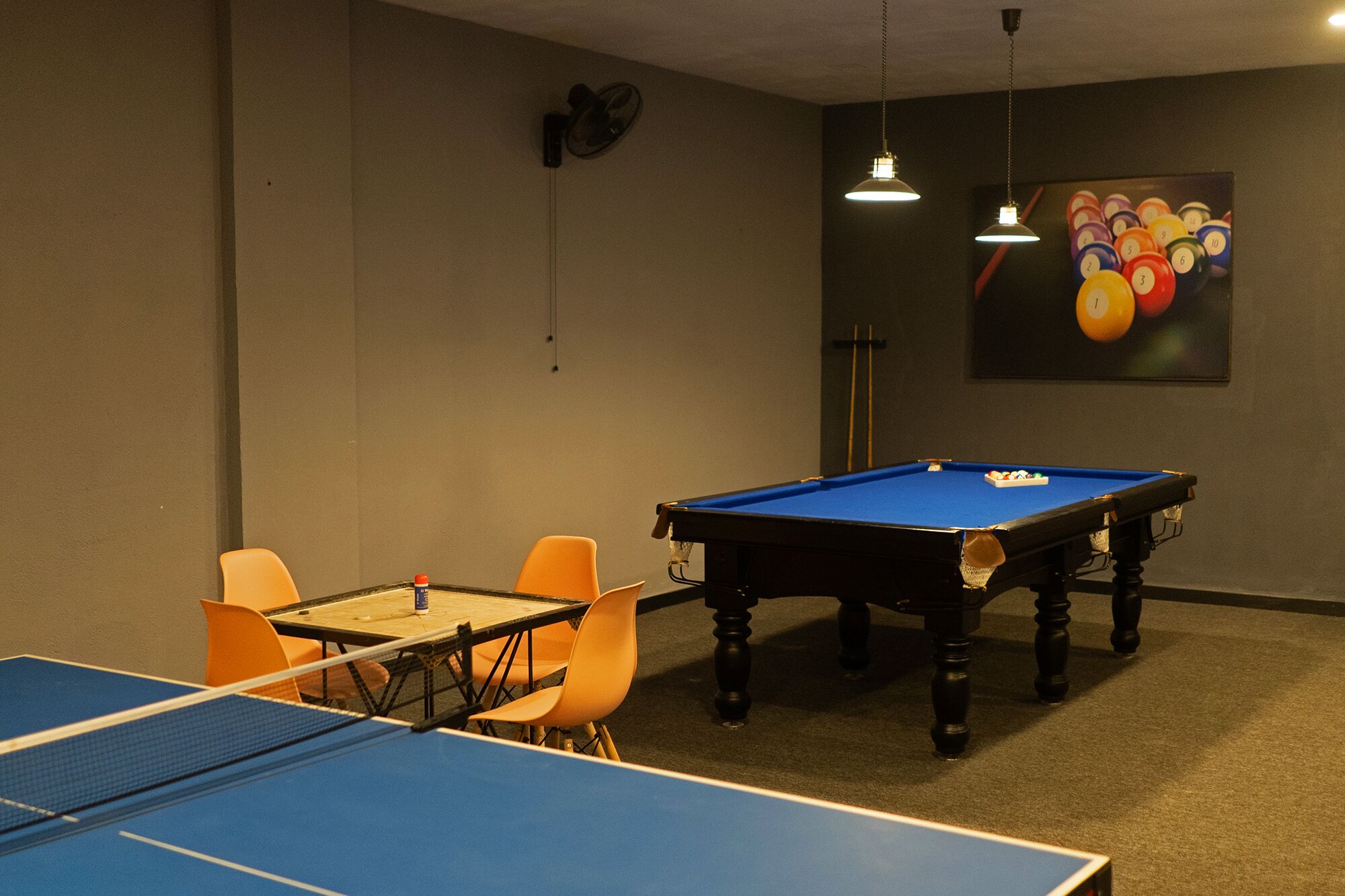 A pool table with orange chairs, a ping pong table, and a billiards table in a room with beige walls and a ceiling fan.