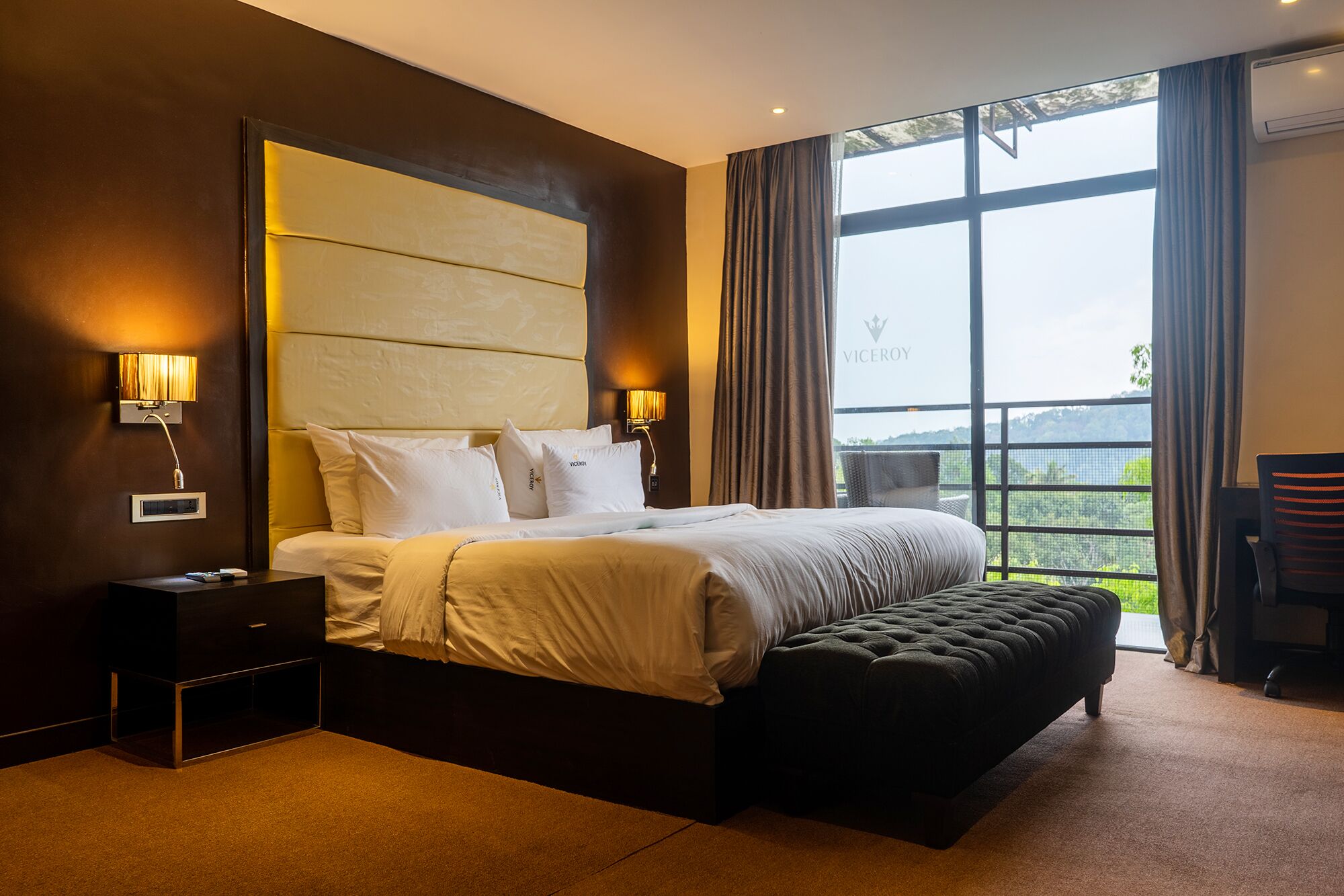 Una habitación de hotel moderna con una cama grande, una mesita de noche, una lámpara de lectura y vistas al exterior a través de un ventanal amplio.