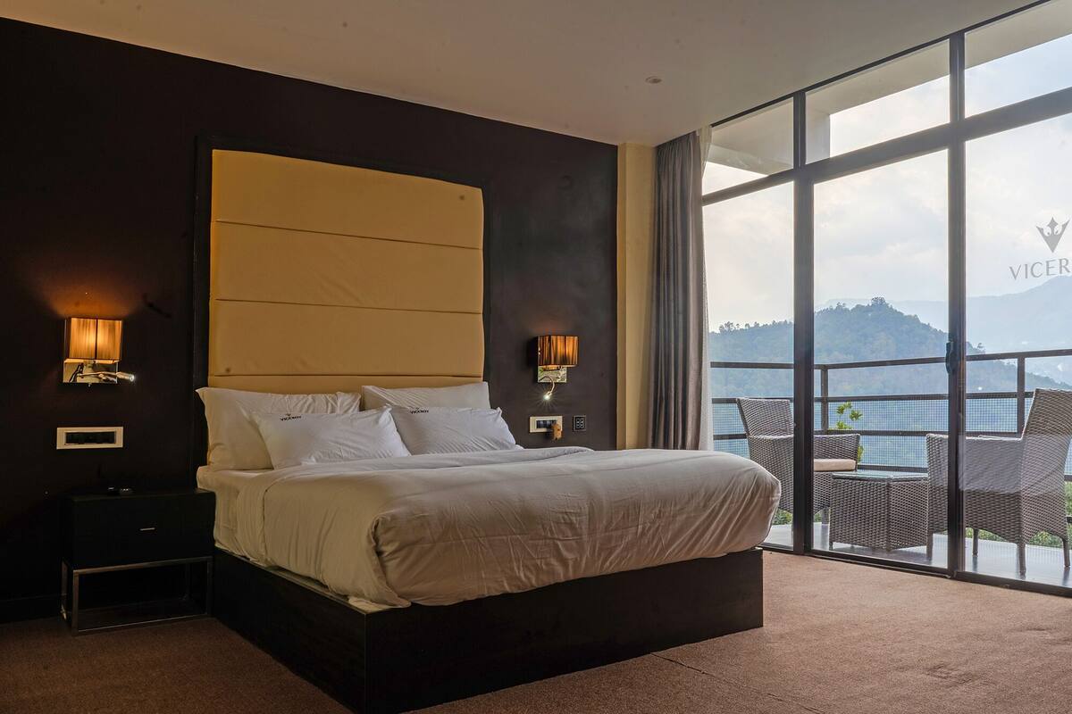 Un dormitorio con una cama grande, dos lámparas de pared, una mesita de noche y vistas a un balcón con muebles de mimbre.