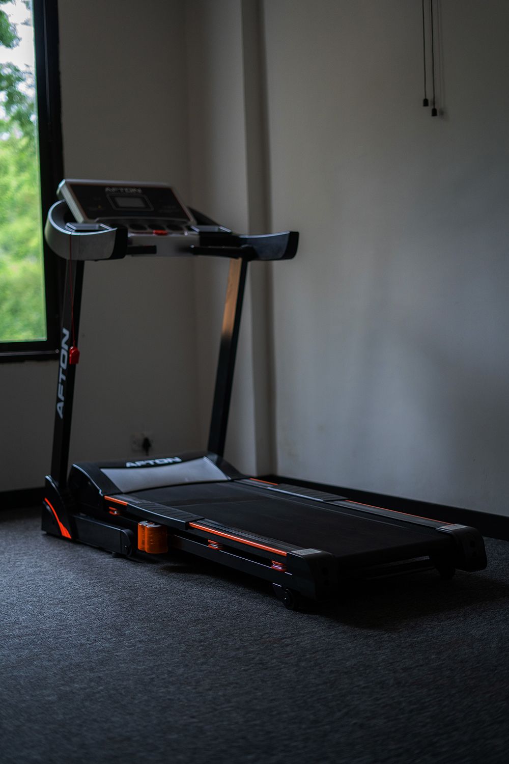 A treadmill in a room with a window, carpeted floor, and a plain wall.