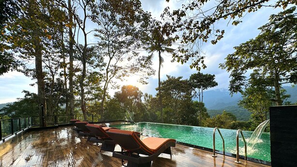 A wooden deck with lounge chairs overlooking a pool, surrounded by trees and a mountainous landscape. - Viceroy Luxury Mountain Resort (Udumbanchola)