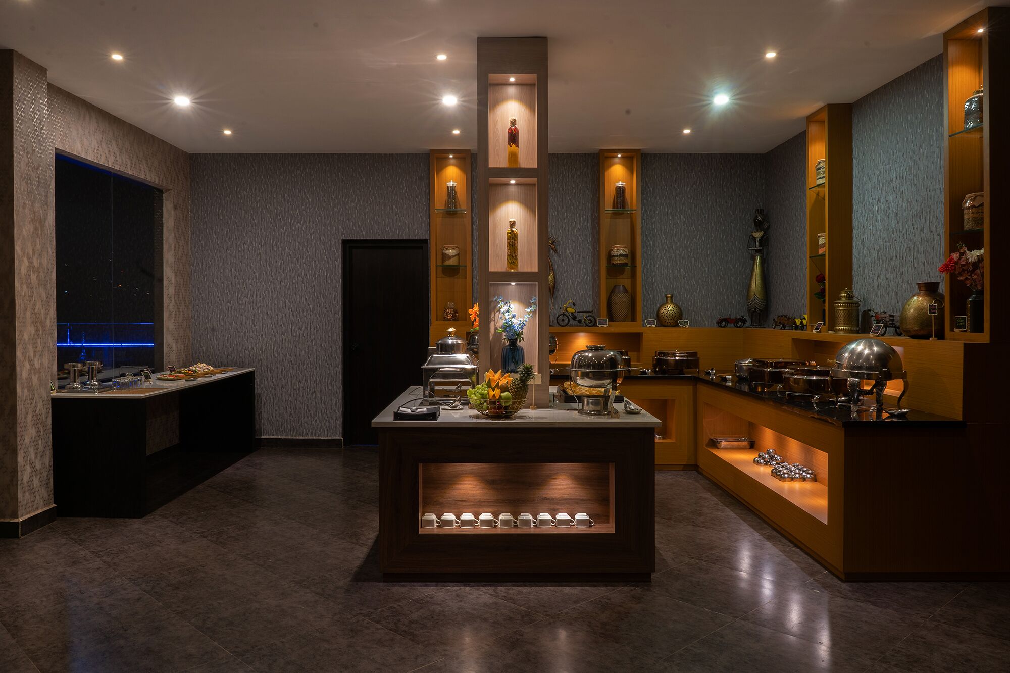 A modern hotel lobby with a central island featuring a coffee station, surrounded by shelves with decorative items and a large mirror.