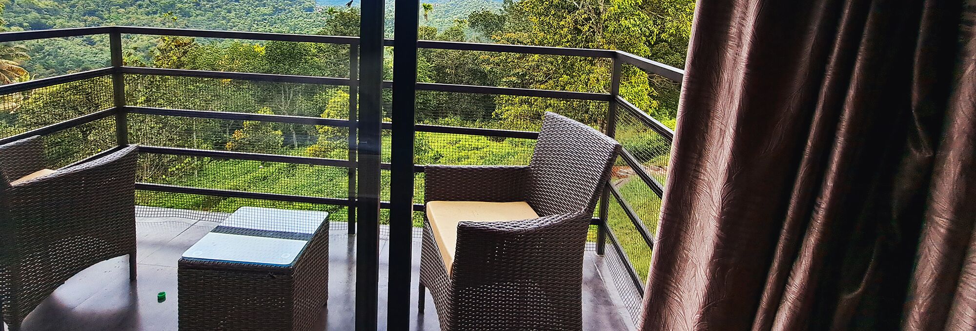 Ein Balkon mit Korbmöbeln und einer Glasbrüstung, mit Blick auf eine üppige grüne Landschaft.