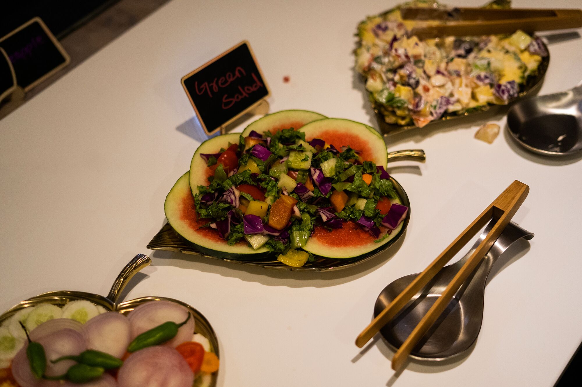 A variety of dishes including a salad in a watermelon bowl, a fruit salad, and a green salad with a sign indicating 'Green Salad'.