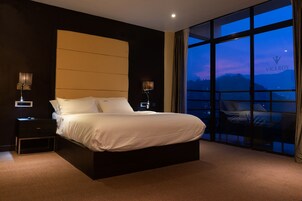 Una habitación de hotel moderna con una cama grande, dos mesitas de noche con lámparas y vistas a las montañas a través de amplias ventanas.