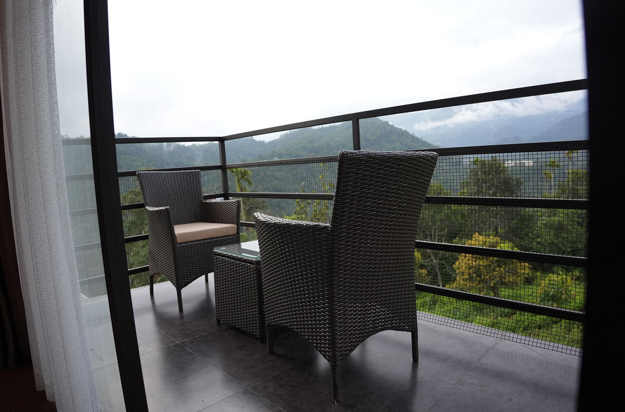 A balcony with wicker furniture and a glass table, overlooking a mountainous landscape.
