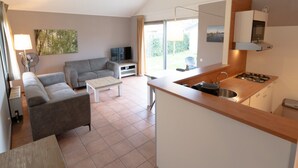 Ein modernes Wohnzimmer mit einer Couch, einem Couchtisch und einem Fernseher. Es gibt eine Küchenzeile mit einem Herd und Schränken.