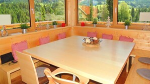 A wooden cabin with a large dining table, red and white checkered pillows, and large windows offering a view of a forested mountain area. - Huttenzauber 1 Comfortable Holiday Residence (Annaberg-Lungoetz)