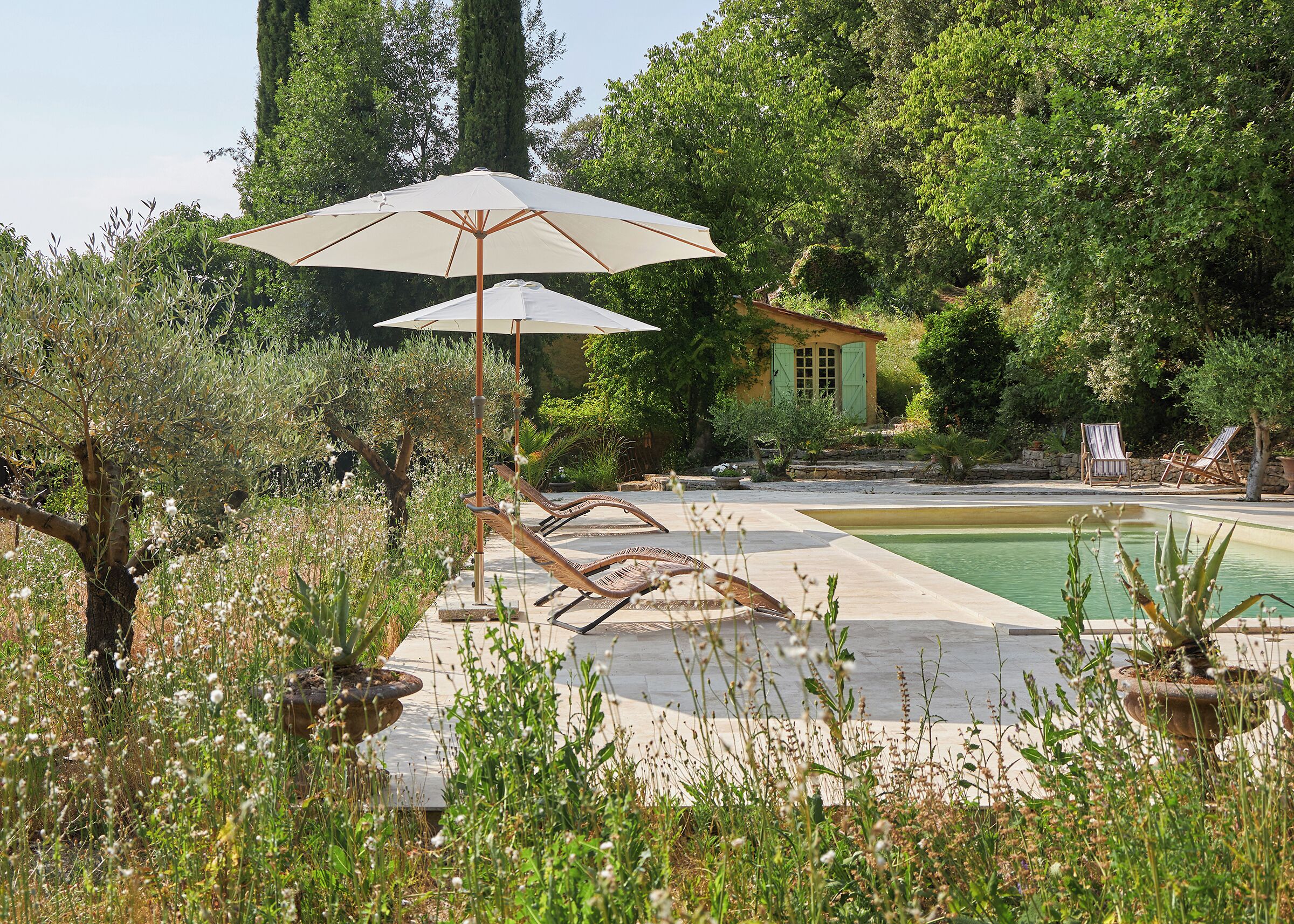 A pool area with lounge chairs and umbrellas, surrounded by greenery and a small building.