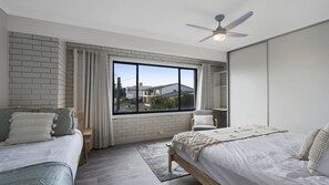 A modern bedroom with two beds, a large window, and a ceiling fan. - Clear Water at The Entrance by HolidayCo. (THE ENTRANCE NORTH)