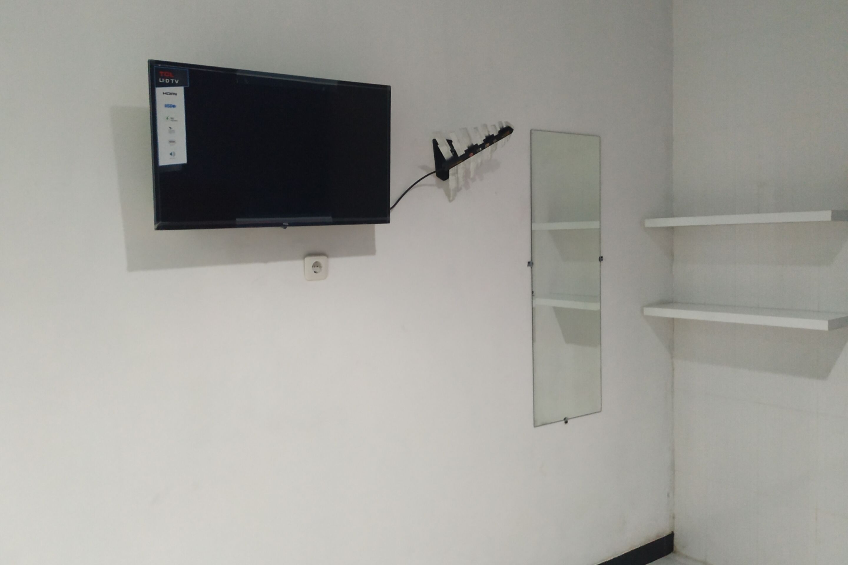 A room with a mounted television, a mirror, and two white shelves.