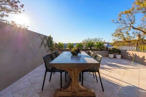 Un espace de restauration en plein air avec une longue table et des chaises, entouré de plantes en pot et offrant une vue sur le paysage urbain.