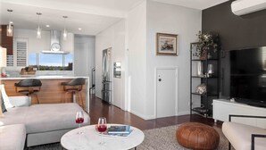 Una sala moderna con sofá, mesa de centro y televisor. Hay una zona de cocina con refrigerador, estufa y taburetes de bar.