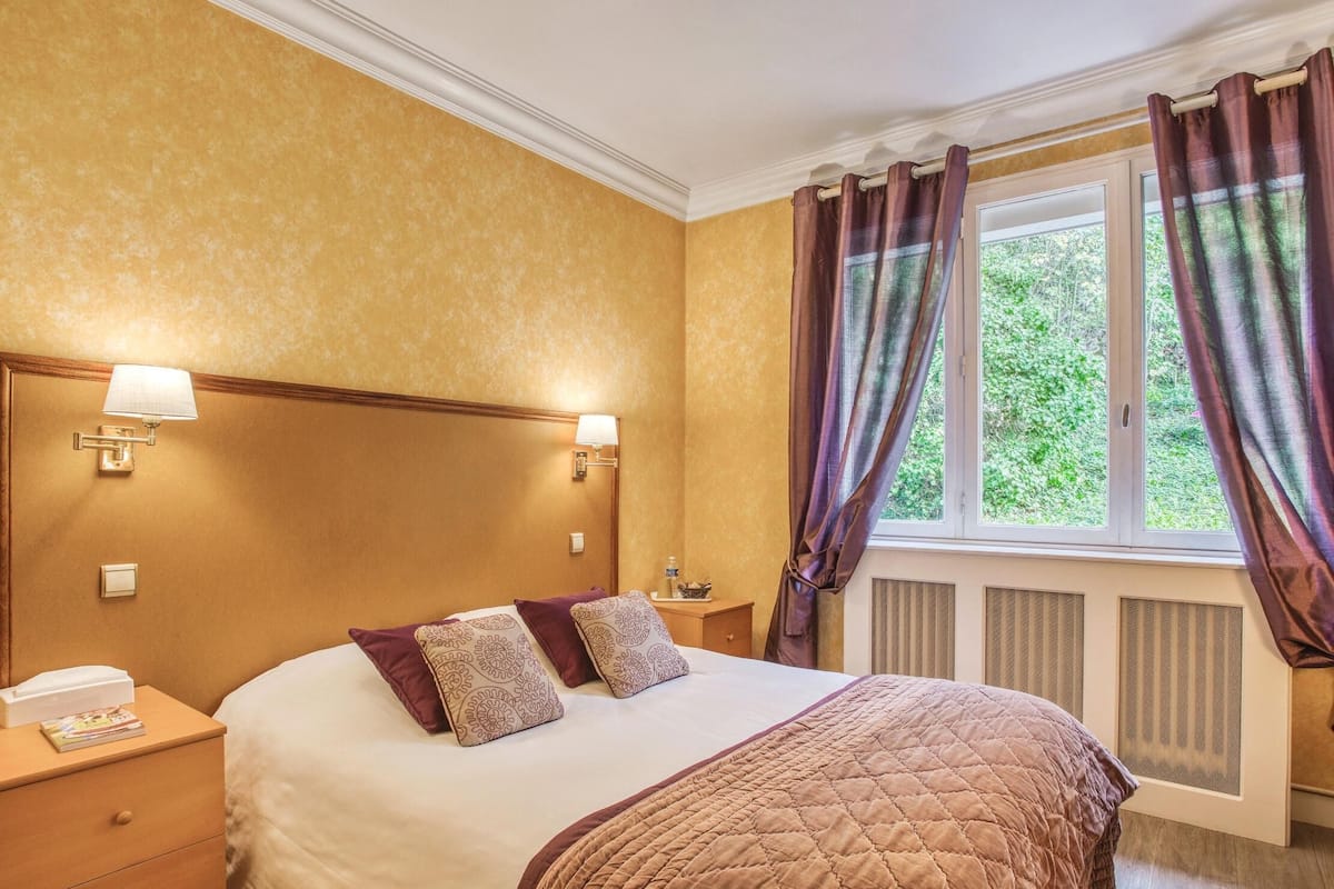 Habitación de hotel con una cama grande, dos lámparas de noche, una mesita de noche y una ventana con cortinas.