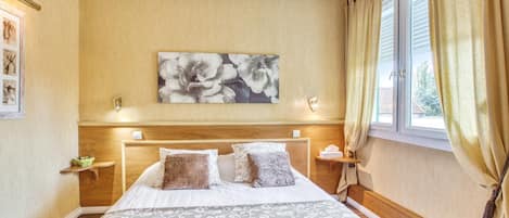 A hotel room with a large bed, a wooden headboard, a floral painting on the wall, and a window with curtains.