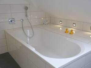 Et moderne badeværelse med et stort, hvidt badekar, et brusehoved og to gummiankerdyr på gulvet.