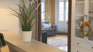 Ein Zimmer mit einem Blumentopf auf einer Holzauslage, einem Schrank mit Glastüren und einem Durchgang zu einem weiteren Raum mit einer Couch und einem Fenster.
