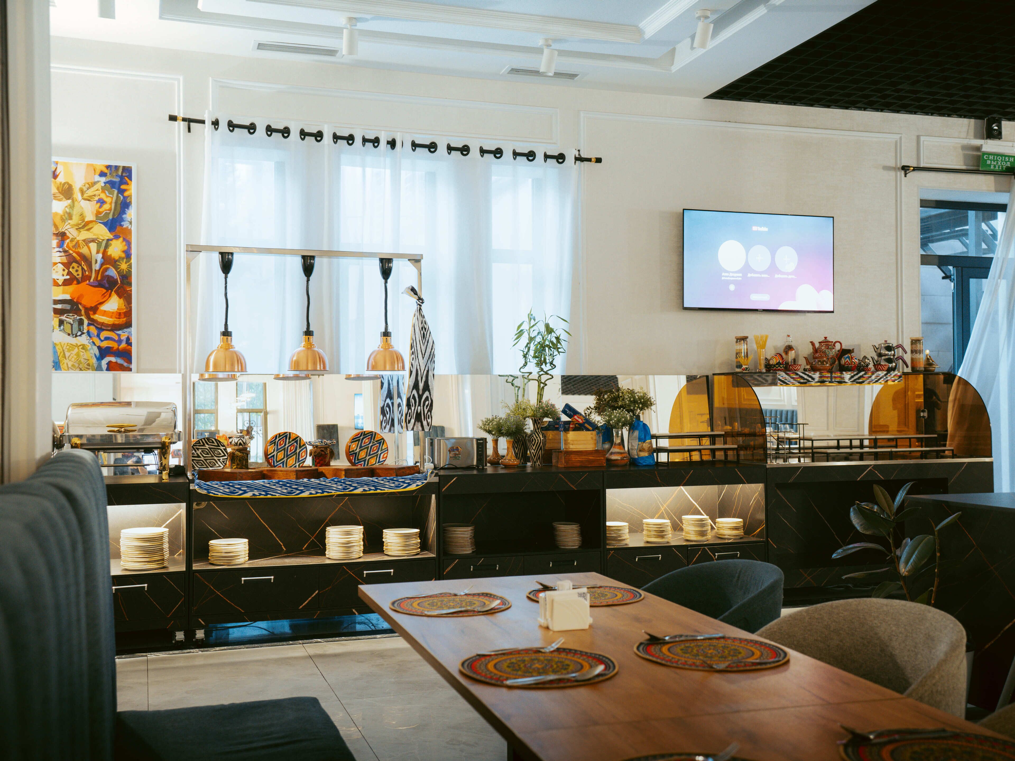 A modern kitchen with a dining area, featuring a long wooden table, hanging pendant lights, and a flat-screen TV mounted on the wall.
