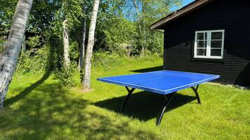 Une table de ping-pong bleue se trouve dans un espace verdoyant, avec des arbres et un chalet en bois en arrière-plan.
