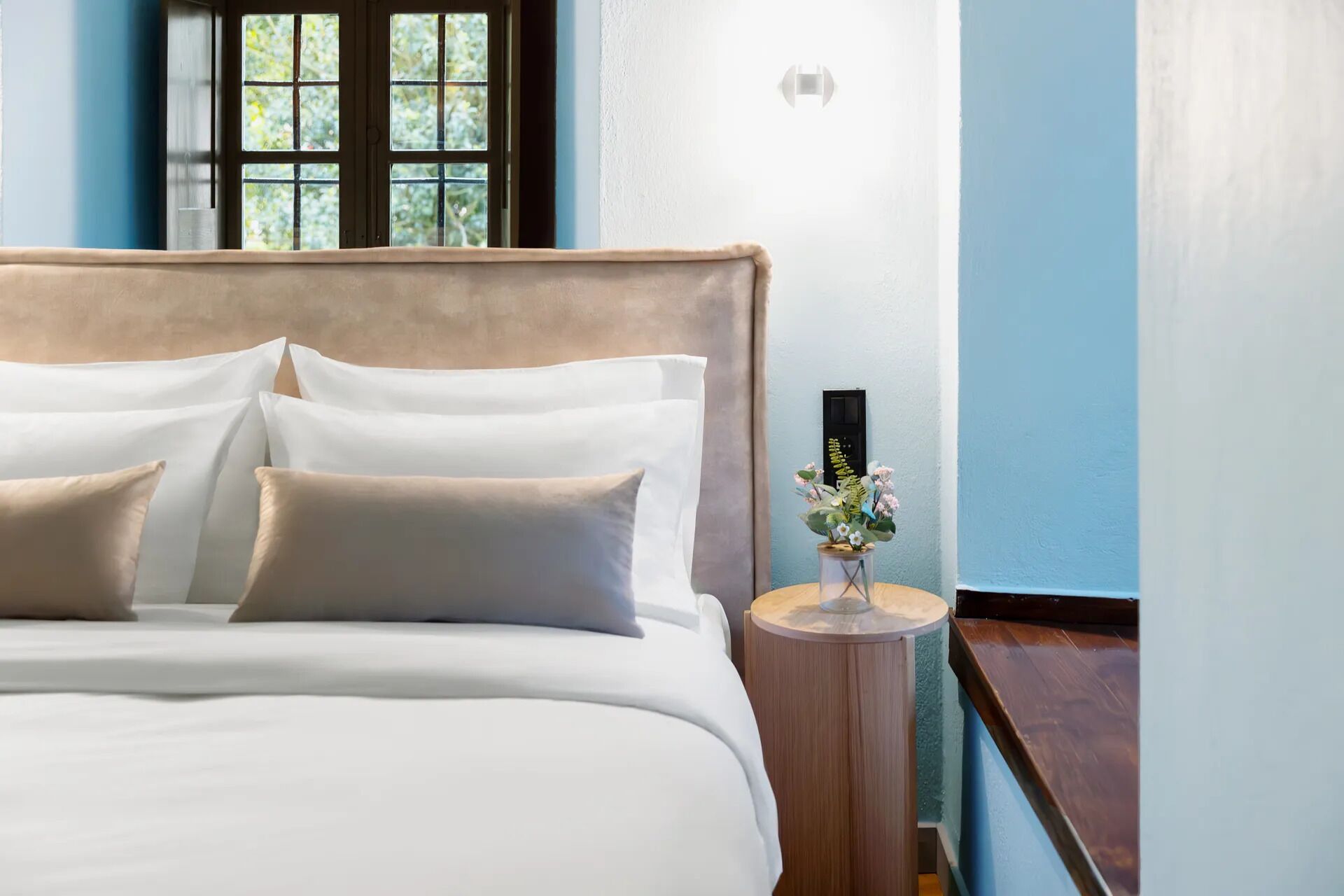 A neatly made bed with white linens and multiple pillows, a small wooden side table with a vase of flowers, and a window with a view of greenery.