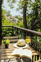 Un banco de madera con un libro abierto, un sombrero de paja, un vaso de líquido amarillo y una pequeña planta en maceta sobre la barandilla de un balcón con vistas a un paisaje verde.